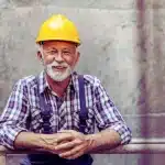 A smiling, senior worker with a helmet on his head, leans on the railing in the factory and takes a break on work. substantial gainful activity exceptions