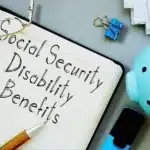 A notebook with the handwritten words "Social Security Disability Benefits," a pen resting on the notebook, a pair of glasses, a blue piggy bank, some paper clips, and a stack of papers. Reduce Disability Benefits